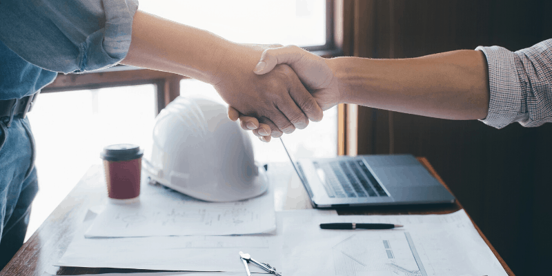 Two adults shake hands across a table with a hard hat and remodeling paperwork.