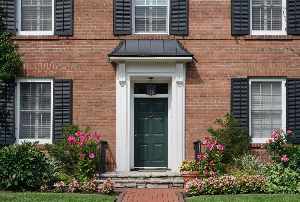 The front of a brick house with a dark green door, black shutters and pink flowers surrounding a small porch.